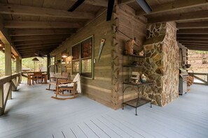 A covered outdoor area with wooden flooring, a seating arrangement including chairs and a table, and a stone wall with a shelf. - Enchanting Creekside Mountain Cabin Retreat with Luxury Amenities in the Heart of Scenic Blue Ridge, Georgia (Blue Ridge)