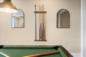 Ein Billardtisch mit grüner Spielvliesbahn, eine Hängeleuchte und zwei an der Wand befestigte Spiegel.