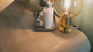 Una mesa de madera con un espejo que refleja una habitación, una lámpara con pantalla de vidrio transparente y un objeto blanco sobre la mesa.