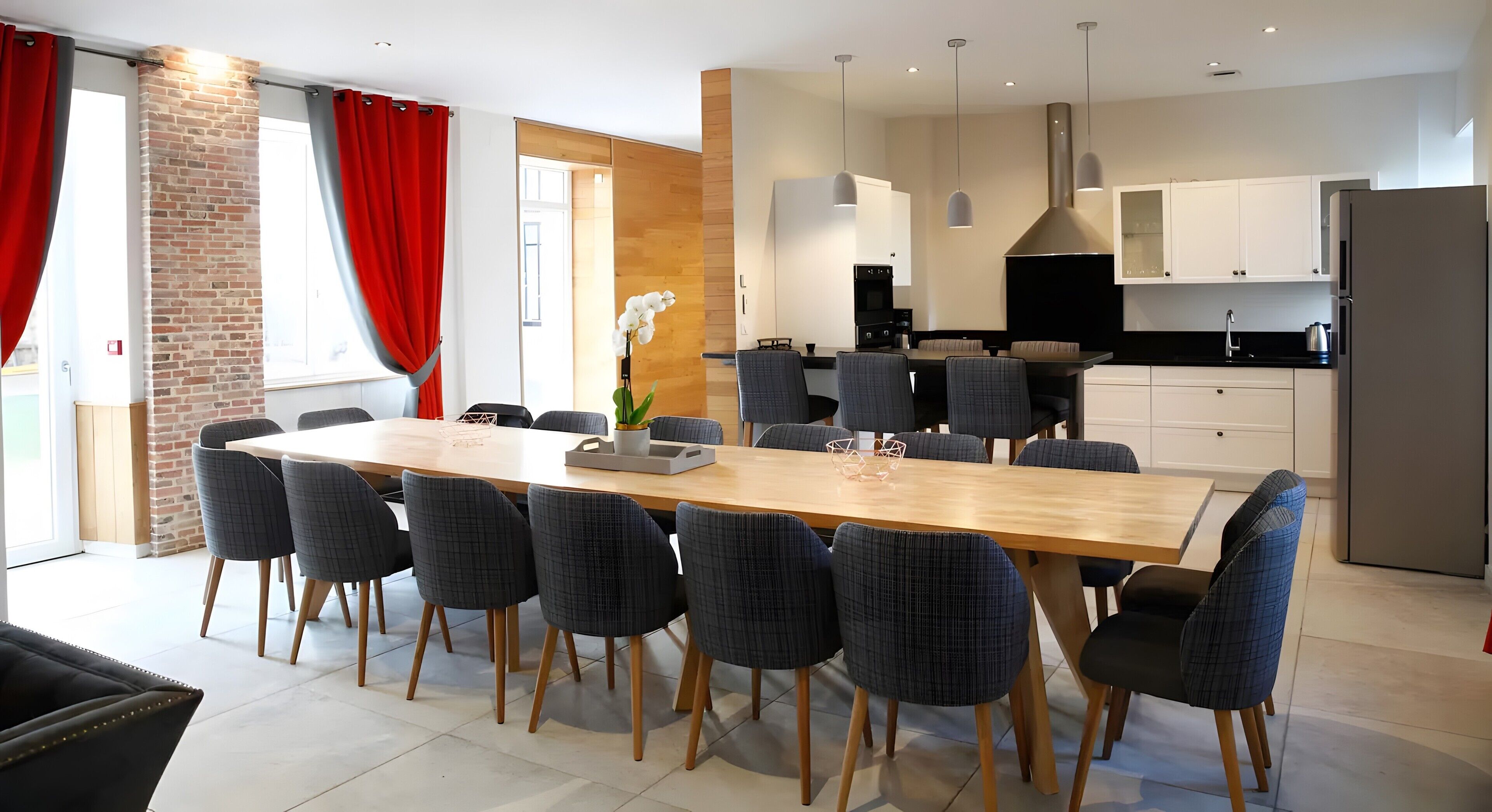 Een moderne eetruimte met een lange tafel en stoelen, een keuken op de achtergrond, en een bakstenen muur.