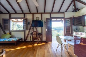 A room with a bed, a desk, a chair, and a table. There is a window with shutters, a clock, and a view of greenery outside. - Bungalow Caféière - Creole Charm in Carbet, Martinique (Le Carbet)