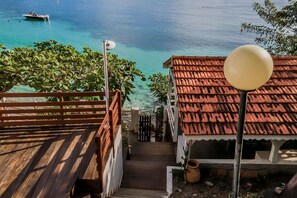 Eine Holzterrasse führt zu einem Gebäude mit rotem Ziegeldach, mit Blick auf ein klares, blaues Meer und ein Boot.