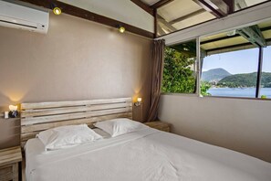 A bedroom with a wooden headboard, two bedside lamps, a window with a view of a body of water and hills, and an air conditioning unit. - Villa Fleur D'eau - Right on the Water (Les Anses-d`Arlet)