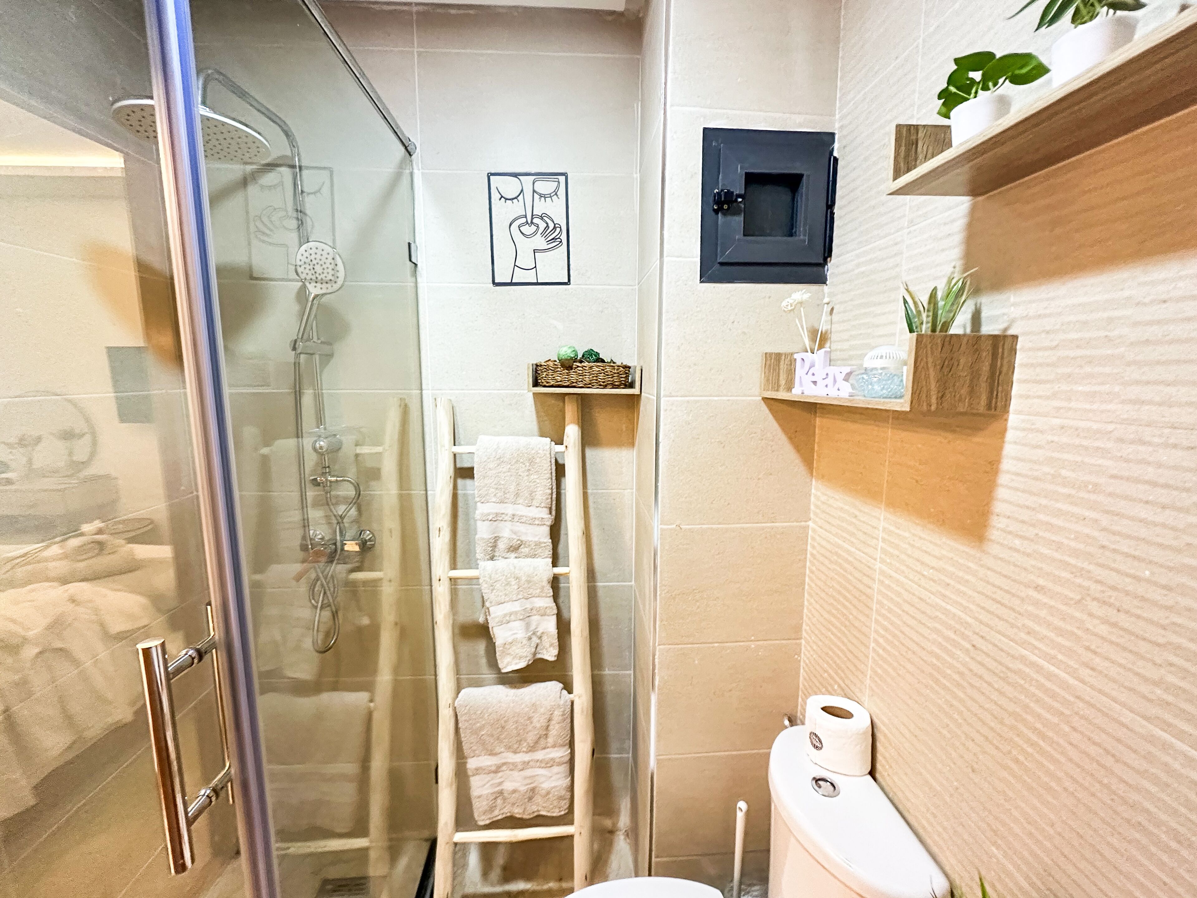 Une salle de bain avec une douche à parois de verre, une échelle en bois sur laquelle sont posées des serviettes, des toilettes et une étagère contenant des articles de toilette.