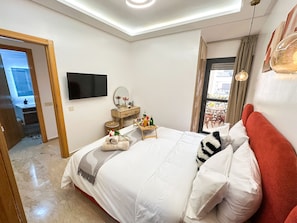 A modern bedroom with a large bed, a flat-screen TV, a bedside table with a mirror, and a view of the balcony.