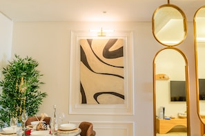 A dining area with a table set for two, a wall-mounted abstract painting, and a mirror with a gold frame.
