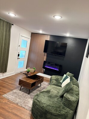 A modern living room with a green sofa, a wooden coffee table, a flat-screen TV mounted on a black wall, and a window with curtains. - Cozy 1BR - Sleeps 4 - Walk to Downtown & Free Parking (Dallas)