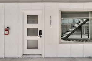 Eine weiße Tür mit drei Glasscheiben und einem Zahlenschloss, ein an der Wand befestigter Feuerlöscher und ein großes Fenster mit Blick auf eine Treppe.