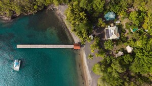 Eine Luftaufnahme eines Küsteneigentums mit einem Steg, der sich in türkisfarbenes Wasser erstreckt, umgeben von dichter Vegetation und einigen Gebäuden.