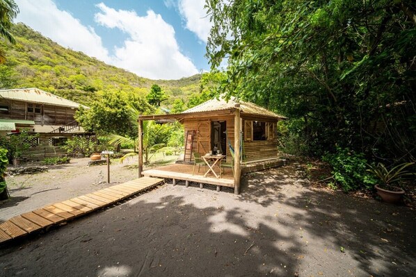 A rustic wooden cabin with a porch, surrounded by lush greenery and a mountain backdrop.