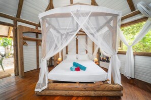 A room with a bed under a canopy, wooden beams, and a view of greenery outside.