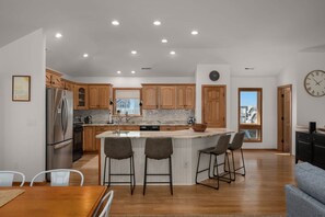 A modern kitchen with wooden cabinets, a stainless steel refrigerator, a central island with bar stools, and a view through the window. - Salty Moon Outer Banks (Currituck County)