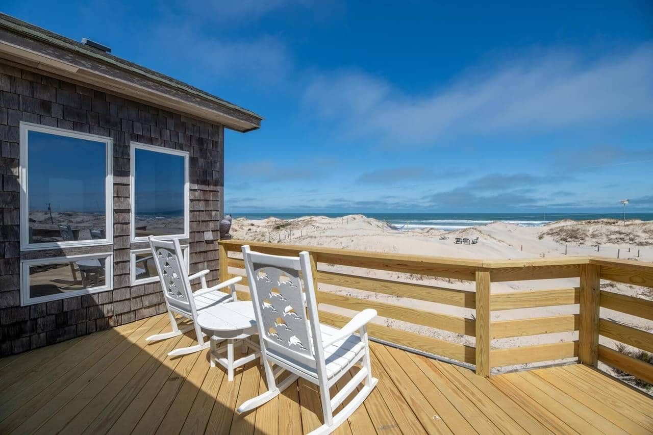 A wooden deck with two rocking chairs and a table, overlooking a beach with sand dunes and the ocean.
