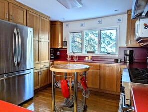 A kitchen with wooden cabinets, a stainless steel refrigerator, a round table with wine glasses, and a window with a view of trees. - Idyllic Waterfront Cottage with Fire Pit Near Acadia National Park | Gouldsboro, Maine (Gouldsboro)