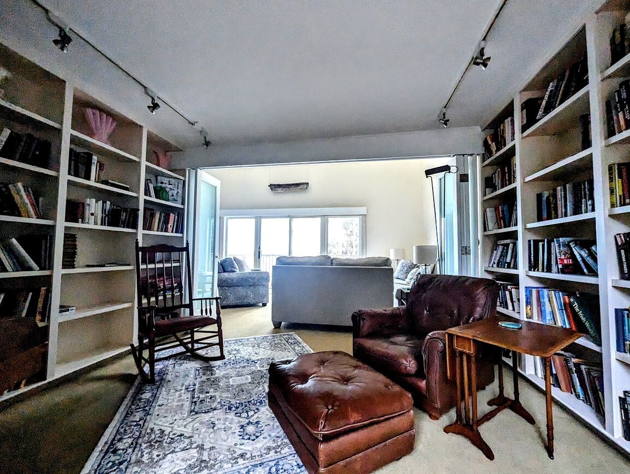 Ein Wohnzimmer mit einer Couch, einem Ledersessel, einem Holztisch und einem gemusterten Teppich. An beiden Seiten des Raumes befinden sich Bücherregale.