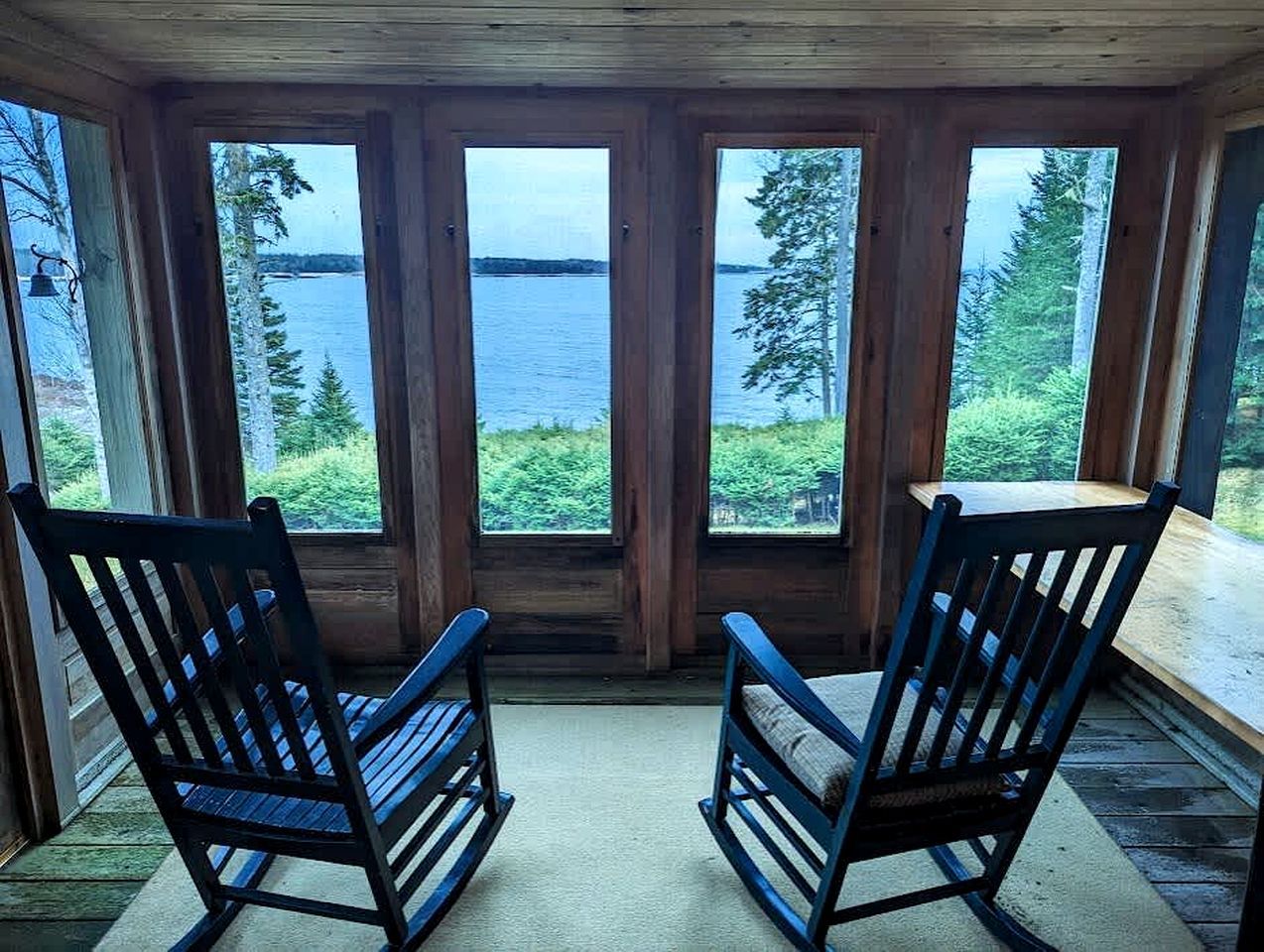 A cabin with a large windowed porch overlooking a lake, two rocking chairs, and a wooden ceiling.