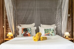 A bed with white linens, two pillows with tropical prints, a yellow towel, and two bedside lamps.
