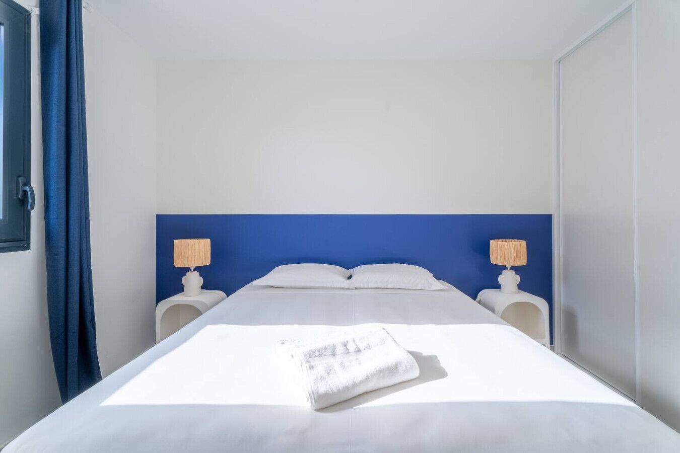 Hotellihuone, jossa on sänky, kaksi yöpöytää, sininen pääty ja verho.