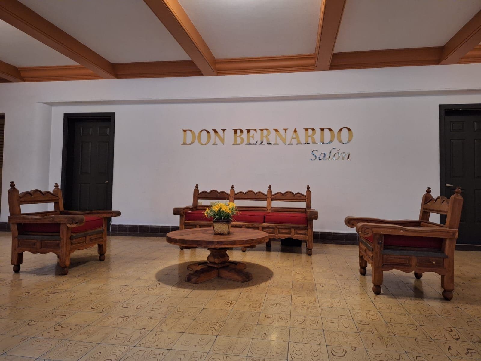Una habitación con un banco de madera, dos sillas y una mesa redonda con un florero. En la pared hay un letrero que dice 'DON BERNARDO Salón'.