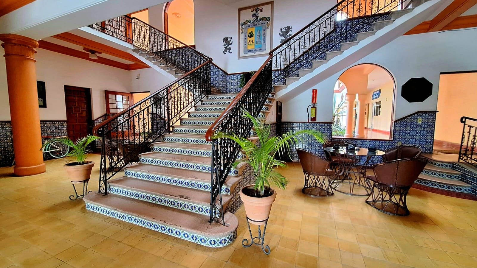Un magnifique escalier, agrémenté de rampes décoratives, mène à un niveau supérieur. Des plantes en pot sont disposées à sa base, et un espace détente avec des chaises et une table se trouve à proximité.