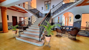 Una imponente escalera con barandales ornamentales que conduce a un nivel superior. Hay plantas en macetas en la base y un área de descanso con sillas y una mesa.