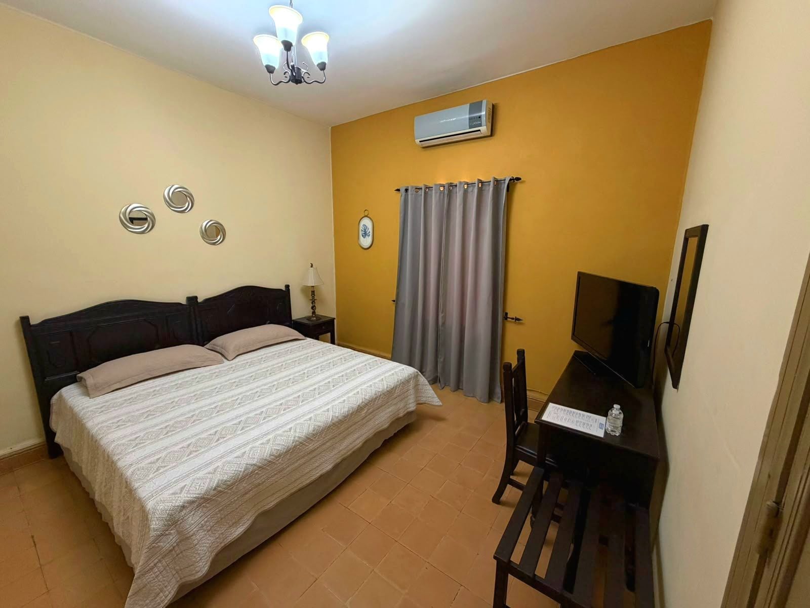 A bedroom with a bed, a desk, a chair, a television, an air conditioning unit, and a curtain.
