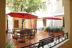 Un patio al aire libre con mesas y sillas, sombrillas rojas y vista a un edificio con ventanas y una puerta.