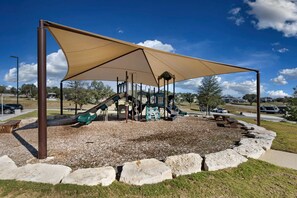 Ein Spielplatz mit Rutsche und Klettergerüsten, umgeben von schattigen Bäumen und einer Steinsicht.