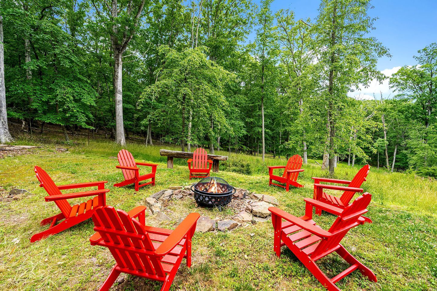 Een vuurkuil omringd door rode Adirondack-stoelen in een gazon met bomen op de achtergrond.