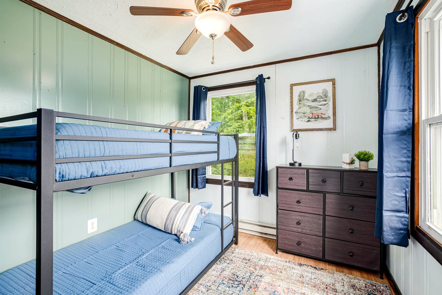 A bunk bed room with a ceiling fan, a dresser, a window with curtains, and a framed picture on the wall.