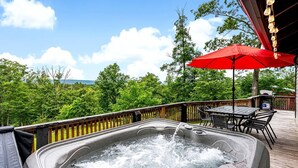 Un jacuzzi en una terraza de madera con una sombrilla roja, rodeado de follaje y un cielo despejado.