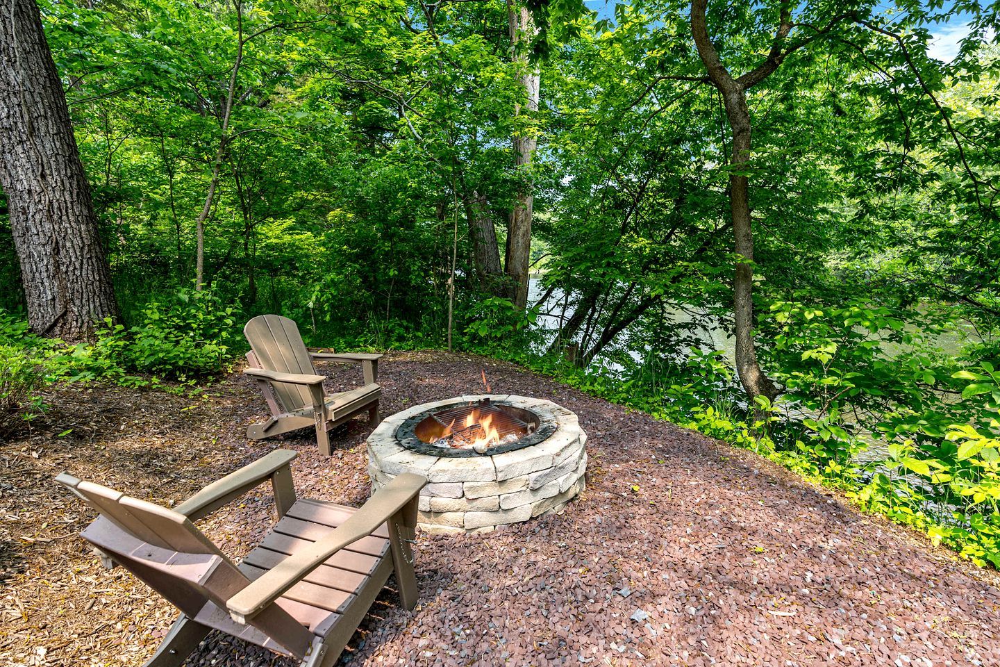 An outdoor seating area with Adirondack chairs and a stone fire pit surrounded by a wooded landscape.