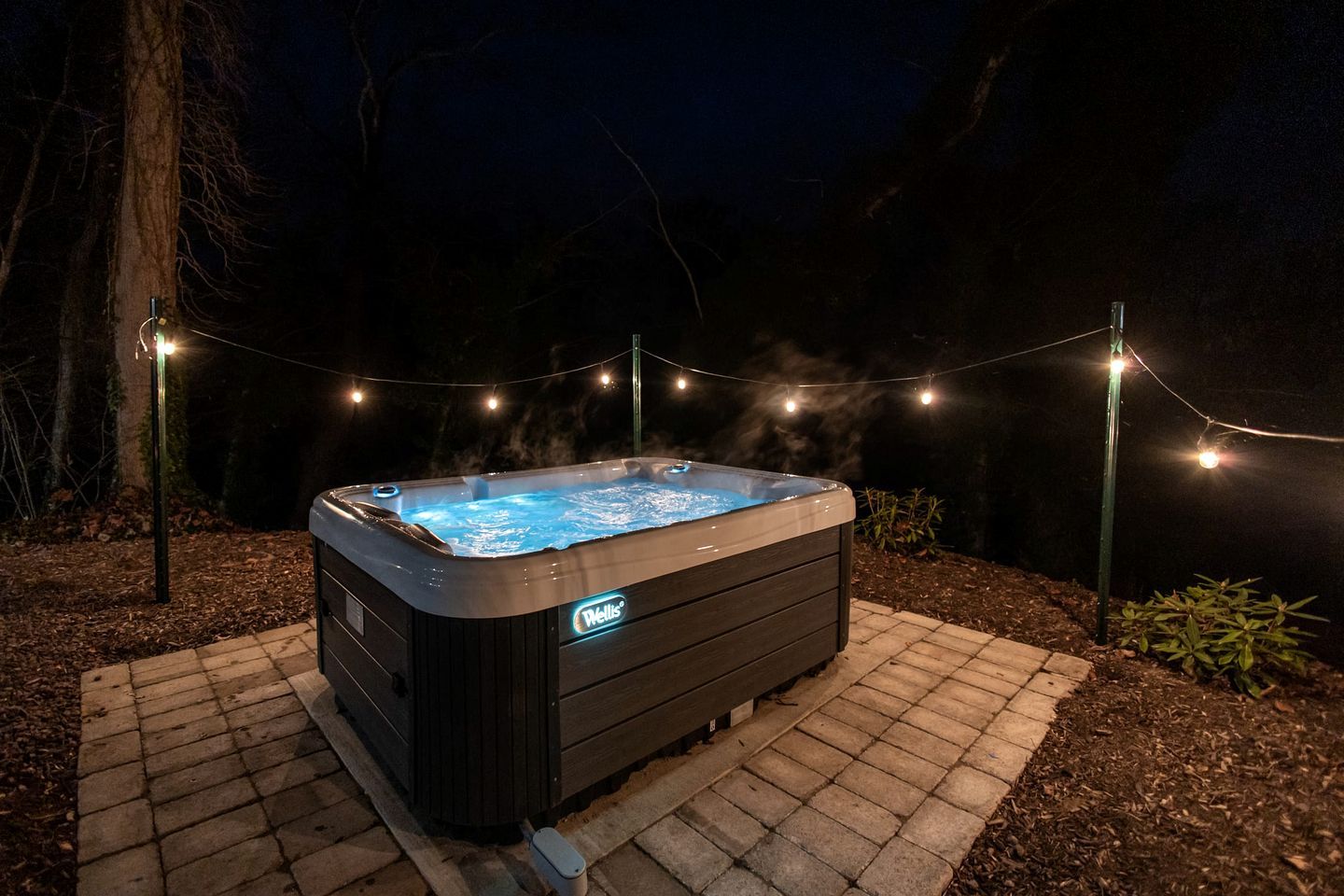 A hot tub with a visible brand logo, surrounded by a string of lights and a paved area.