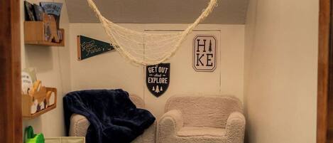 Une pièce chaleureuse avec un hamac, une chaise, un tapis et une étagère remplie de livres et ornée d’une pancarte.