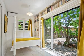 A bedroom with a bed, a bookshelf, a window with curtains, and a view of the outside. - Les Beaux Arts Bungalow (Le Diamant)