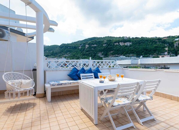 Una terraza en la azotea con una mesa blanca dispuesta, una silla colgante y un banco blanco con cojines azules.