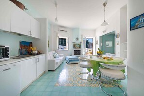 Une cuisine moderne avec des meubles blancs, un coin repas doté d’une table verte et un salon avec un canapé et une vue sur l’extérieur.