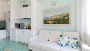 Un salón con sofá blanco, un cuadro de un pueblo costero y una zona de cocina con armarios y electrodomésticos blancos.