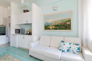 Ein Wohnzimmer mit weißem Sofa, einem Gemälde einer Küstenstadt und einer Küchenzeile mit weißen Schränken und Geräten.