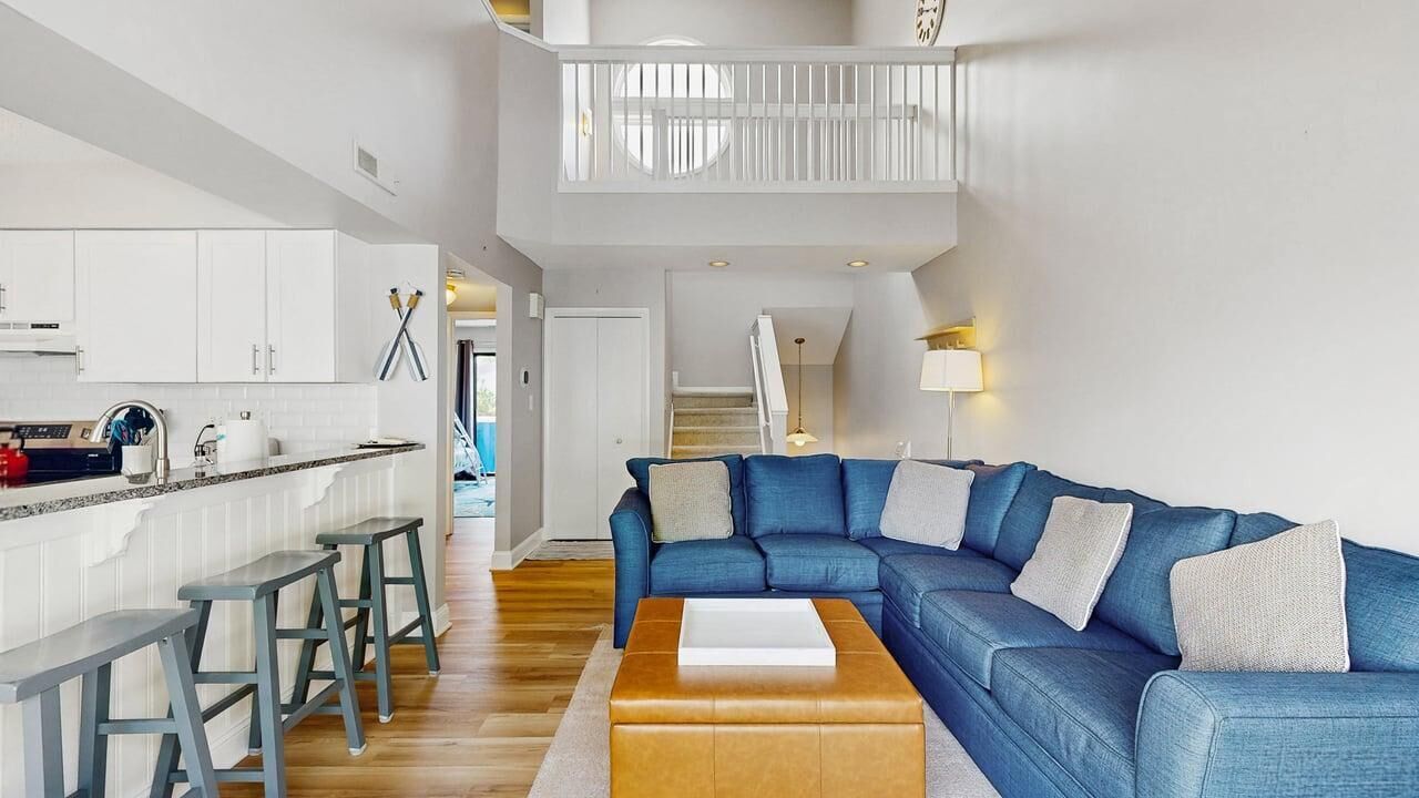 A modern living room with a blue sofa, wooden coffee table, and a kitchen area with white cabinets and a bar stool.
