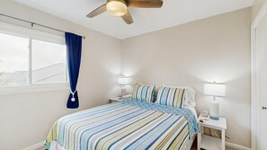 Ein Schlafzimmer mit einem Bett, Nachttischlampen, einem Deckenventilator, einem Fenster mit Vorhängen und Blick ins Freie.