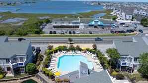 A residential area with a large swimming pool surrounded by lounge chairs and umbrellas, a parking lot, and a view of the sea. - East of the Sun - 39916 E Sun Dr #426 (Fenwick Island)