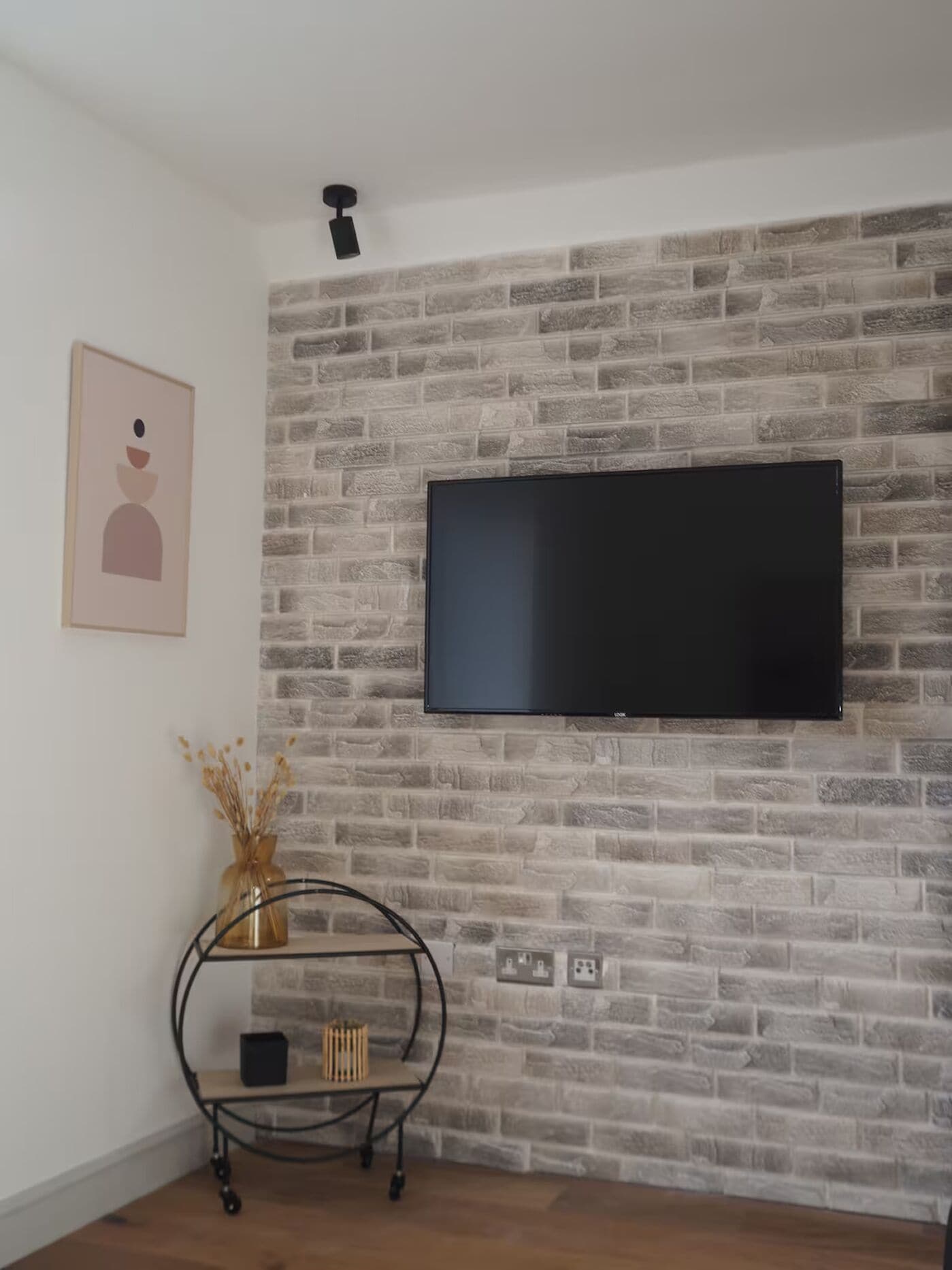 A room with a brick-patterned wall, a mounted flat-screen TV, a small shelf with decorative items, and a power outlet.