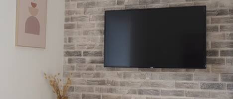 A room with a brick-patterned wall, a mounted flat-screen TV, a small shelf with decorative items, and a power outlet.