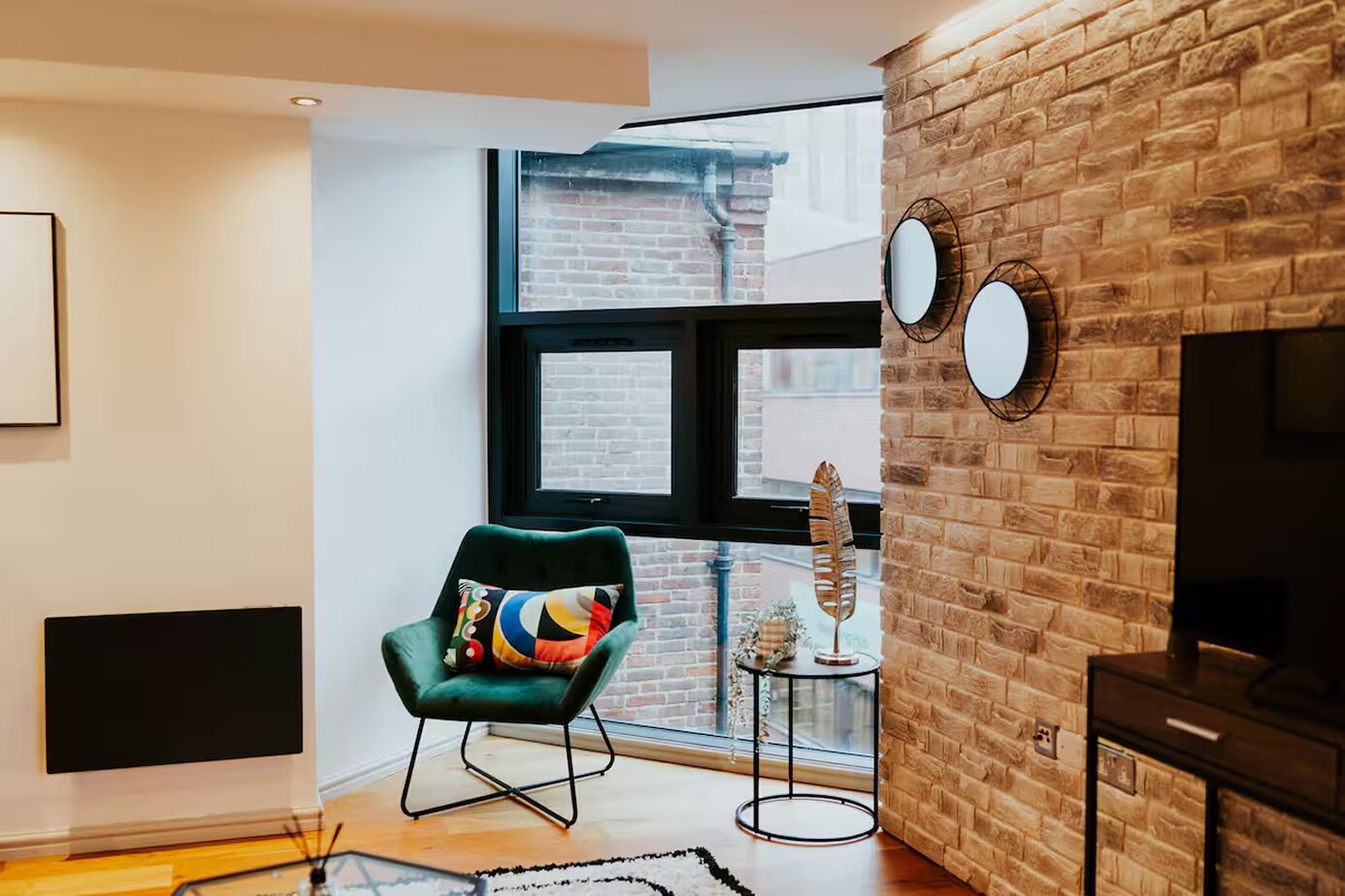 A modern living room with a brick wall, a large window, a green armchair, a small side table, and a flat-screen TV.