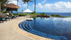A pool with a curved edge, lounge chairs, a thatched umbrella, and a view of the ocean. - Princeville One Bedroom Villa Resort w/ Amenities (Princeville)