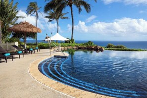 A pool with a curved edge, lounge chairs, a thatched umbrella, and a view of the ocean. - Princeville One Bedroom Villa Resort w/ Amenities (Princeville)