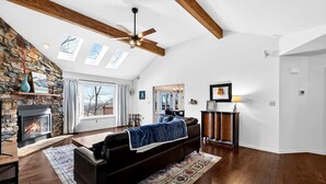 A spacious living room with a fireplace, a ceiling fan, and a large window. - Stunning Hedgesville Mountain Home with Fire Pit & Screened Porch Near Hedgesville, WV (Hedgesville)