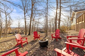 Eine Gruppe roter Adirondack-Stühle ist um eine Feuerstelle in einer bewaldeten Außenanlage angeordnet, mit einem Haus im Hintergrund.