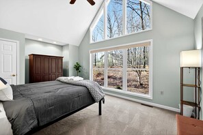 Quarto com uma cama grande, um guarda-roupa, uma janela com vista para árvores e um ventilador de teto.